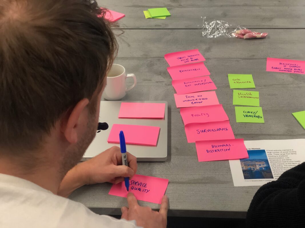 Over-the-shoulder view of a person engaged in a brainstorming session. The wooden table is covered with pink and green sticky notes containing various written concepts. The person is writing 'Service Quality' on a pink note with a blue marker. A pink mug is visible, along with a laptop partially in frame. The notes include topics like 'Surveillance', 'Poverty', and 'Health instrument'. The scene suggests a collaborative planning or problem-solving exercise in progress.