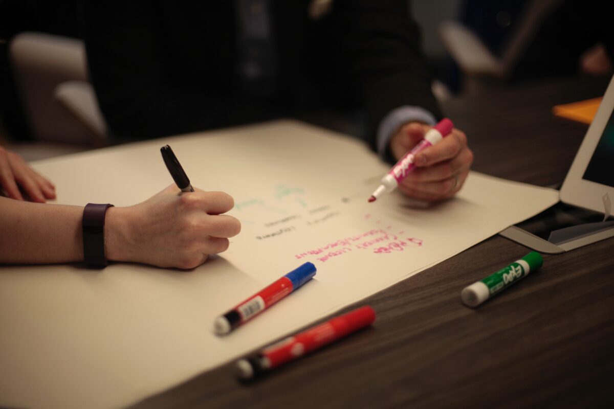 Close-up of a table with hands visible, engaged in collaborative work. One hand holds a black pen over a large sheet of paper, while another holds a pink marker. Various colored markers are scattered on the wooden table surface. A partial view of a laptop is visible. The image conveys a sense of brainstorming or planning session in progress.