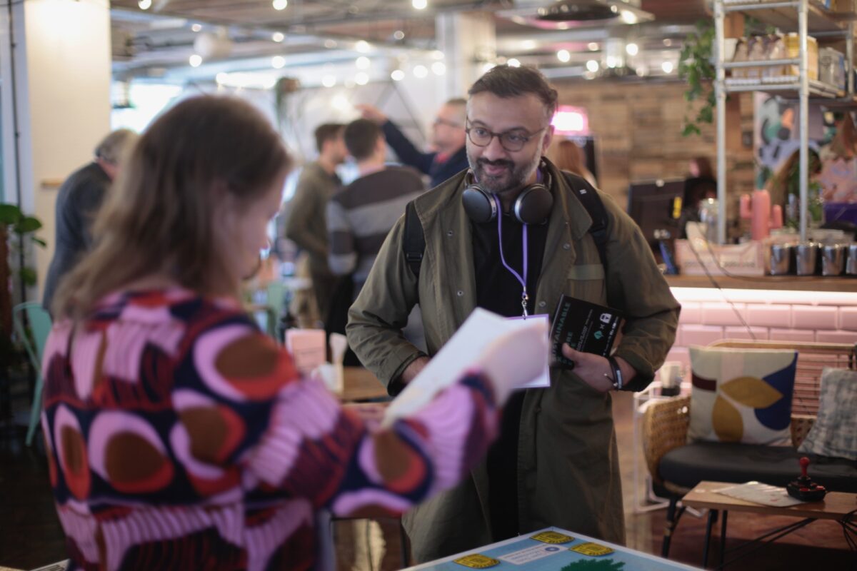 A close-up of two people interacting at what appears to be a conference or event. In the foreground, a woman wearing a colorful patterned top is holding some papers. Facing her is a bearded man with glasses, wearing a green jacket and headphones around his neck. He's smiling and also holding papers and a device. The background shows a busy, well-lit indoor space with other people and event materials visible.